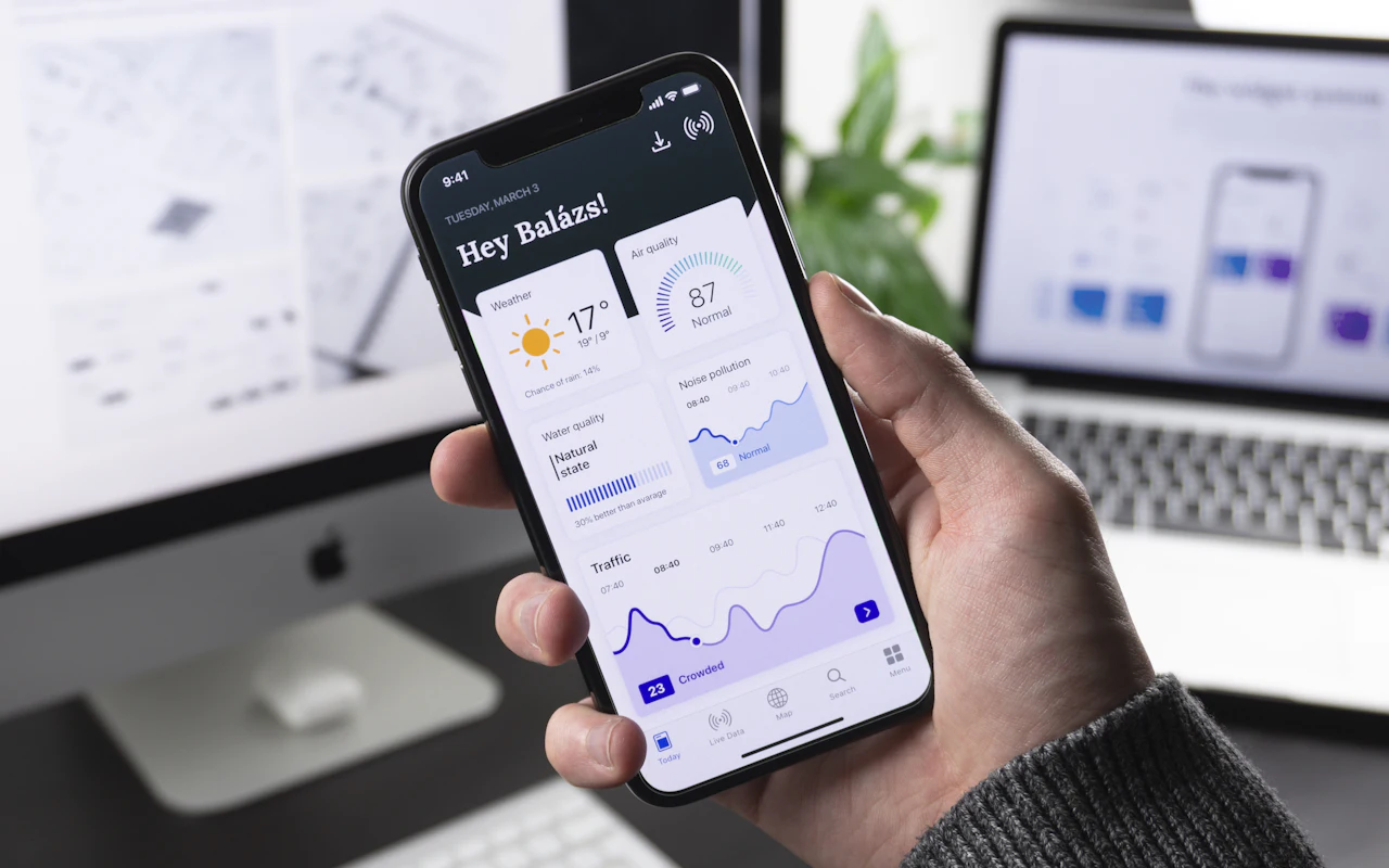 Ruka drží telefon s aplikací a grafy, v pozadí notebook s návrhem obrazovek.